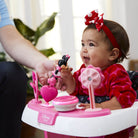 Baby in a high chair with pink toys and a red bow, interacting with an adult hand.