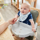Baby sitting in a high chair with a bib, holding a hand, in a home setting.