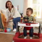 Child in a red and white walker with a woman sitting on a couch in a living room.